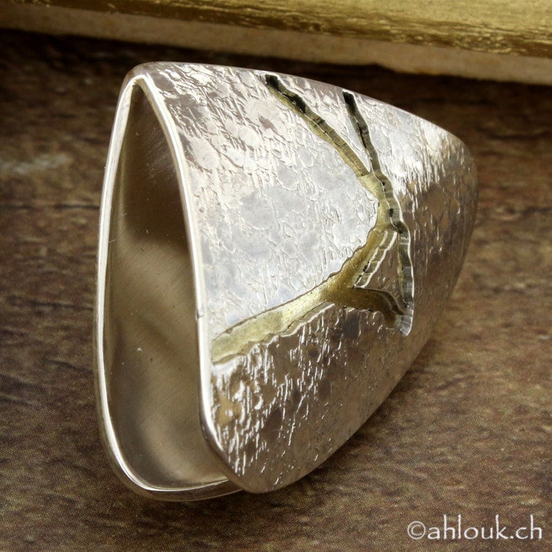 May include: A silver ring with a textured surface and a gold-colored accent. The ring has a rectangular shape with rounded edges and a split design. The gold accent resembles a tree branch.