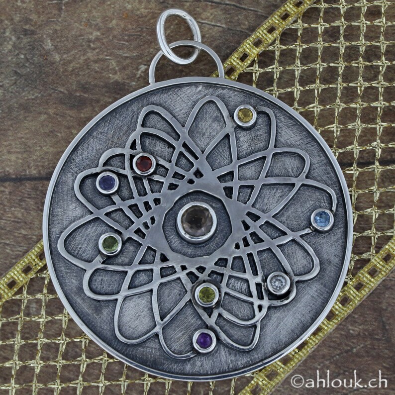 May include: A silver pendant with a circular design featuring a central gemstone surrounded by smaller gemstones in various colors. The pendant has a textured surface and a delicate, intricate pattern.
