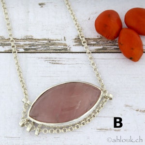 Puede incluir: Un collar de cadena de plata con un colgante de cuarzo rosa. El colgante tiene forma ovalada y est&aacute; engastado en un bisel de plata. El collar est&aacute; sobre una superficie de madera blanca con bayas rojas en el fondo. La letra "B" es visible en la esquina inferior derecha de la imagen.