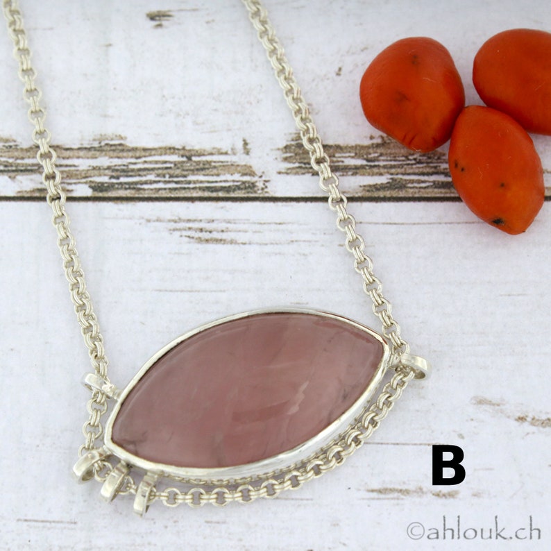 Puede incluir: Un collar de cadena de plata con un colgante de cuarzo rosa. El colgante tiene forma ovalada y est&aacute; engastado en un bisel de plata. El collar est&aacute; sobre una superficie de madera blanca con bayas rojas en el fondo. La letra "B" es visible en la esquina inferior derecha de la imagen.