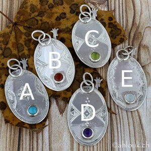 May include: Five silver oval pendants with intricate etched designs and gemstone accents. Each pendant features a different colored gemstone: turquoise, red, green, purple, and light blue. The pendants are displayed on a wooden surface with a leaf.