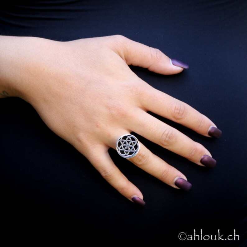 May include: A silver ring with a geometric design. The ring is worn on the middle finger of a hand with dark nail polish.