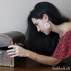 May include: A woman with long black hair is looking at a wooden treasure chest with metal accents. She is wearing a red and white floral dress, silver earrings, and a silver bracelet.