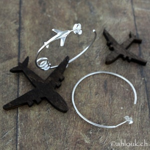 May include: A pair of silver earrings, one with a small airplane charm and the other a simple hoop. The earrings are displayed on a wooden surface.