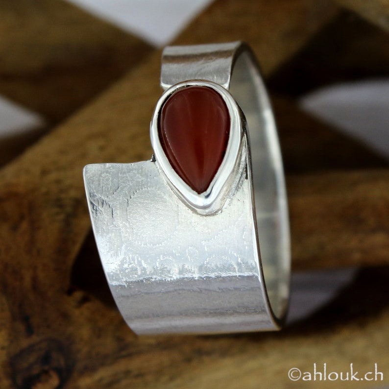 May include: A silver ring with a teardrop-shaped carnelian gemstone set in the center. The ring has a textured, hammered finish.