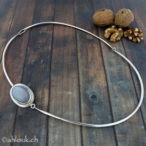 Peut inclure: Un collier ras de cou argent&eacute; avec un pendentif ovale en pierre grise. Le collier est sur une surface en bois.