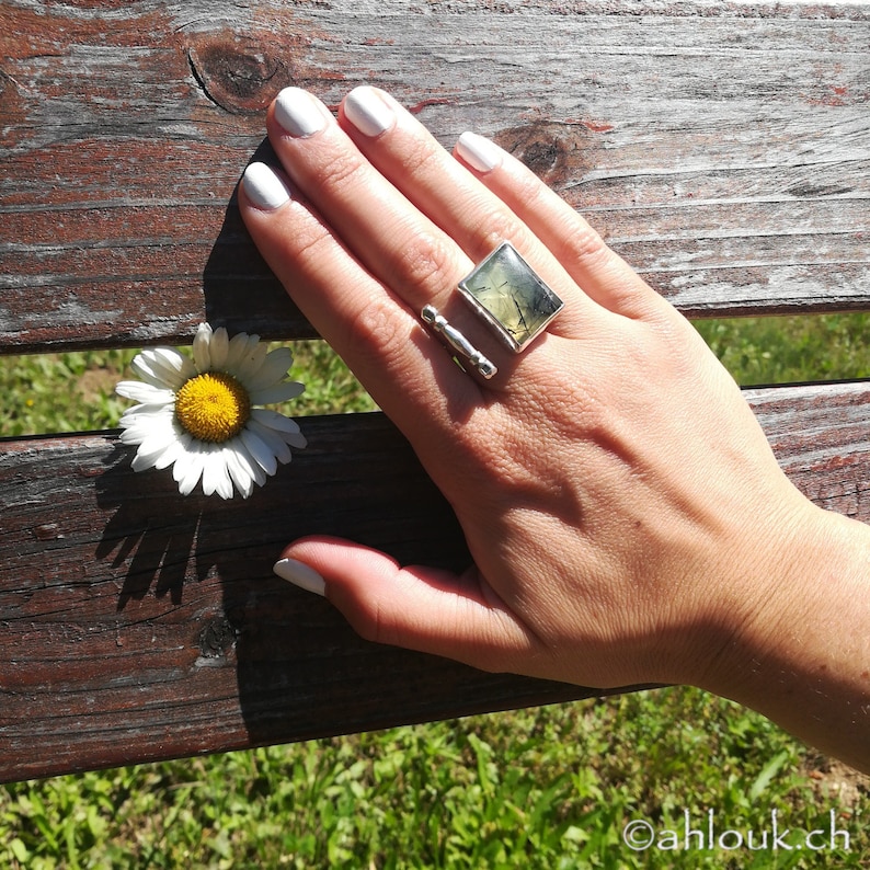 May include: A silver ring with a square green stone set in a silver band. The ring is on a person's finger.