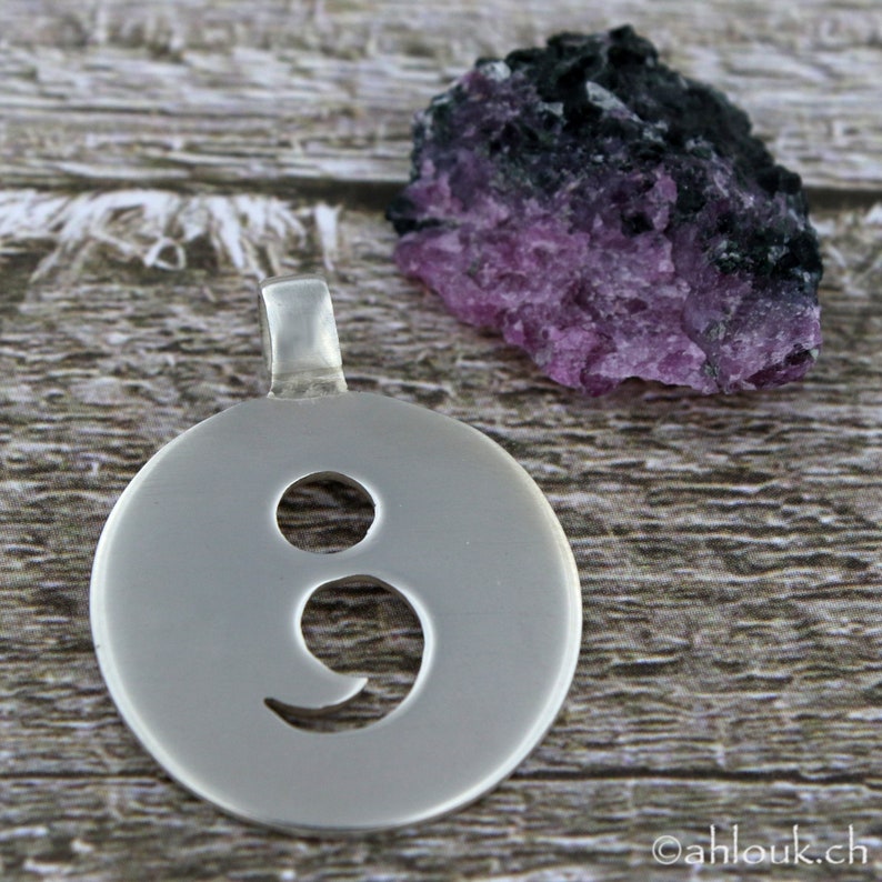 May include: A silver pendant with a semicolon cutout design. The pendant is hanging from a silver chain. The pendant is on a wooden surface.