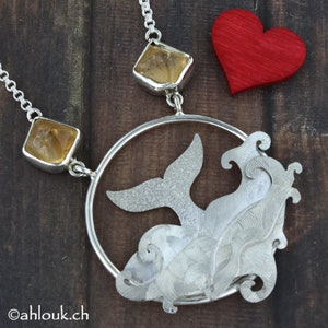 May include: A silver necklace with a whale tail pendant and two amber stones. The whale tail is surrounded by a circle of silver and waves. The necklace is on a wooden background.