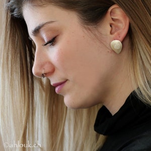 May include: A close-up of a woman's face with a gold earring featuring a rose design. The earring is in the shape of a teardrop. The woman is wearing a black turtleneck sweater.
