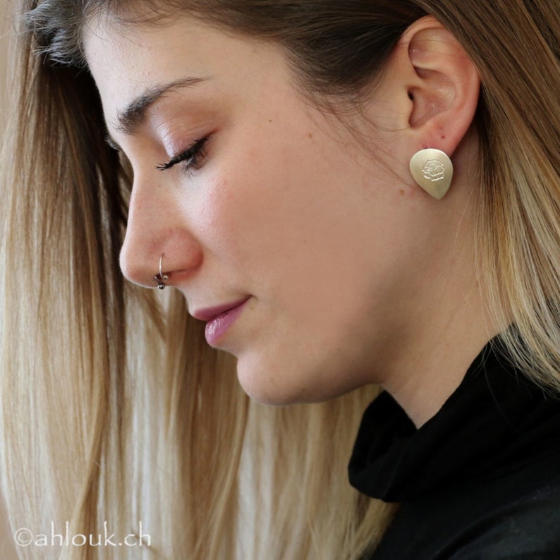 May include: A close-up of a woman's face with a gold earring featuring a rose design. The earring is in the shape of a teardrop. The woman is wearing a black turtleneck sweater.