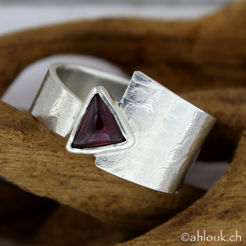 May include: A silver ring with a triangular shaped, dark purple gemstone set in the center. The ring band is textured and has a unique, asymmetrical design.