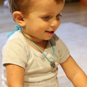 May include: A young child wearing a white t-shirt with a turquoise necklace and a silver pendant. The pendant has a black and white design of a happy face.