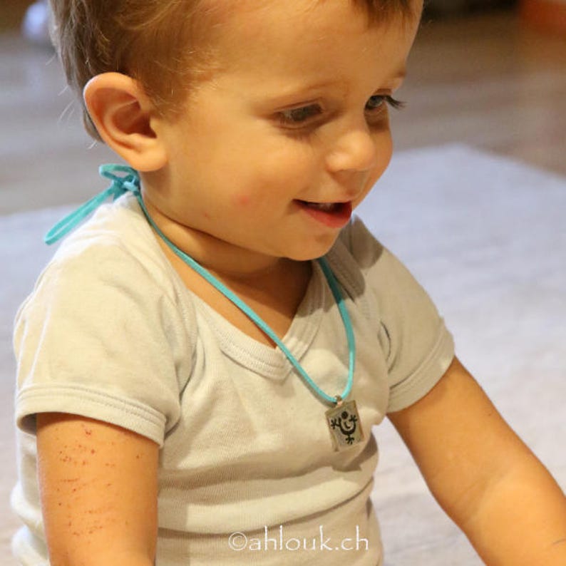May include: A young child wearing a white t-shirt with a turquoise necklace and a silver pendant. The pendant has a black and white design of a happy face.