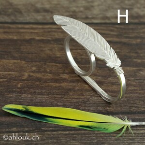 May include: A silver feather ring with a double band design. The ring is made of sterling silver and features a detailed feather design. The ring is shown on a wooden surface with a yellow and green feather.