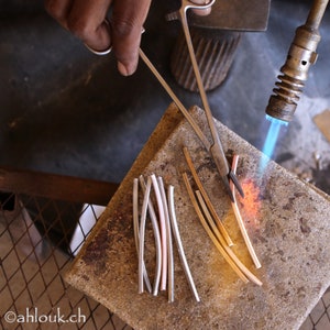 May include: A close-up of a metalworking process, showing a person using a torch to heat metal rods. The rods are arranged on a gray surface, and a pair of pliers is used to hold them in place.