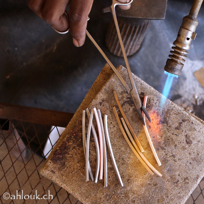 May include: A close-up of a metalworking process, showing a person using a torch to heat metal rods. The rods are arranged on a gray surface, and a pair of pliers is used to hold them in place.