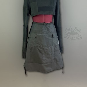 Puede incluir: Una falda utilitaria gris oscuro con múltiples bolsillos y cintura con cordón. La falda se combina con un suéter negro recortado.