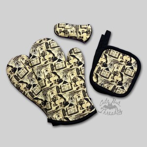 May include: A set of kitchen textiles with a spooky theme. Includes oven mitts, a pot holder, and a bow-shaped item. The items feature a cream-coloured background with a black and white pattern of skulls, tombstones, and insects. Black trim outlines each piece.