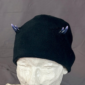 May include: Black fleece hat with two small, curved, blue and white horns. The hat is displayed on a white mannequin head. The horns are a striking contrast to the dark hat.