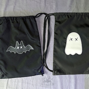 May include: Two black drawstring bags with white embroidered designs. One bag has a bat with cross eyes and the other has a ghost with cross eyes.