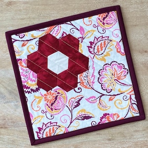 May include: A white quilted fabric coaster with a burgundy floral design. The center of the coaster features a burgundy fabric flower with a white center.