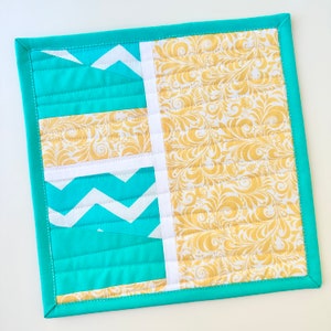 May include: A turquoise blue quilted fabric coaster with a white and yellow geometric pattern. The coaster has a white border and a turquoise blue border.