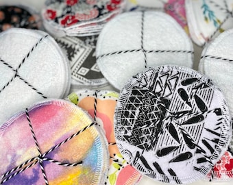 May include: A close-up of a variety of reusable makeup remover rounds. The rounds are made of white cotton fabric and have different patterns printed on them, including floral, geometric, and abstract designs. The rounds are tied together with black string.