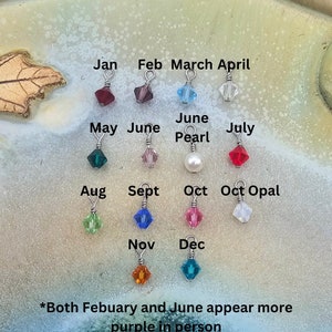 May include: A set of 12 small crystal charms, each representing a different month of the year. The charms are attached to silver rings and are displayed on a light brown surface. The charms are in various colors, including red, blue, green, purple, and white.