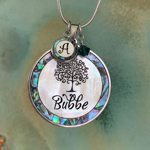 May include: A silver necklace with a round pendant featuring a tree of life design and the word "Bubbe" in cursive. The pendant is made of iridescent abalone shell and has a small silver charm with the letter "A" on it.