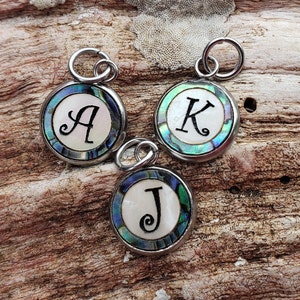 May include: Three silver charms with abalone shell inlays. Each charm features a different letter: A, K, and J. The charms are on a wooden background.