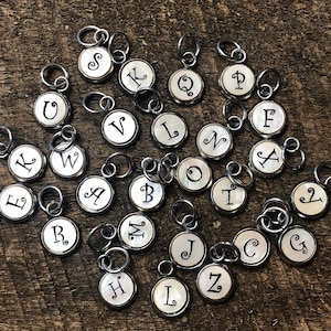 May include: A set of 26 silver charms with a white background and black letter engravings. Each charm features a different letter of the alphabet.