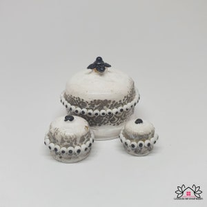 May include: Three white ceramic containers with black and white trim. The largest container has a black knob on the lid. The containers are decorated with a black and white pattern.
