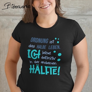 Może przedstawiać: Czarny t-shirt z niemieckim tekstem "ORDNUNG ist das HALBE LEBEN. ICH lebe definitiv in der anderen HÄLFTE!" w kolorze niebieskim i turkusowym. Koszulka ma okrągły dekolt i krótkie rękawy.