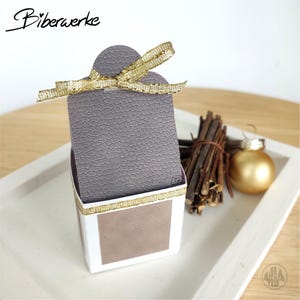 Peut inclure: Une boîte cadeau décorative avec un couvercle gris texturé et un nœud en ruban doré. La boîte a une base blanche avec un panneau marron et une bordure dorée. L'image comprend également une boule dorée et un bouquet de brindilles.