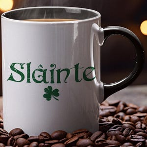 May include: White ceramic coffee mug with a black handle. The mug has a green shamrock and the text "Sláinte" in green lettering. The mug is sitting on a bed of coffee beans.