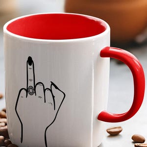 May include: White ceramic coffee mug with a red interior and handle. The mug features a black and white illustration of a hand giving the middle finger with a ring on the middle finger.
