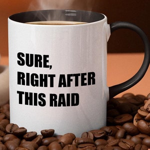 Könnte beinhalten: Eine weiße Kaffeetasse mit schwarzem Rand und Henkel. Die Tasse hat einen schwarzen Schriftzug mit der Aufschrift "SURE, RIGHT AFTER THIS RAID". Die Tasse steht auf einem Bett aus Kaffeebohnen.