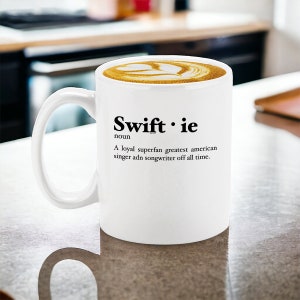 Pu&ograve; includere: Tazza in ceramica bianca con la parola "Swiftie" e una definizione stampata in nero. La tazza contiene un latte con un design decorativo. La definizione recita: "Una superfan leale, la pi&ugrave; grande cantante e cantautrice americana di tutti i tempi."