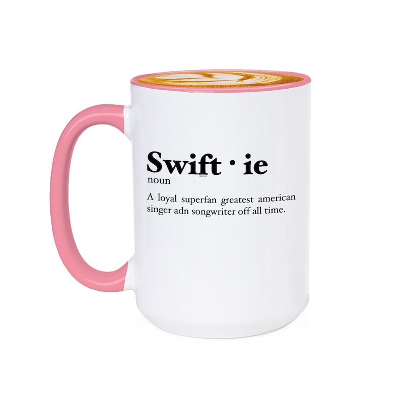 Pu&ograve; includere: Tazza in ceramica bianca con manico e bordo rosa. La tazza presenta la parola "Swiftie" in testo nero, insieme a una definizione. La tazza &egrave; riempita con un latte macchiato, con un disegno sopra.
