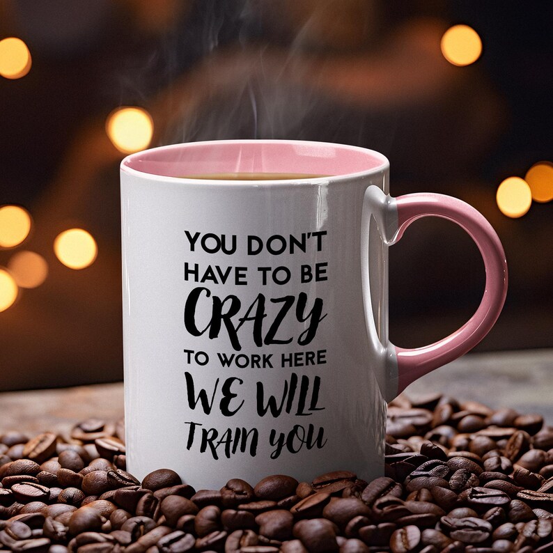 K&ouml;nnte beinhalten: Eine wei&szlig;e Kaffeetasse mit rosafarbenem Rand und Henkel. Die Tasse hat einen schwarzen Schriftzug mit der Aufschrift "You don't have to be crazy to work here, we will train you."