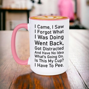 May include: White ceramic coffee mug with a pink rim and handle. The mug has black text that reads: "I Came, I Saw, I Forgot What I Was Doing, Went Back, Got Distracted And Have No Idea What's Going On. Is This My Cup? I Have To Pee."