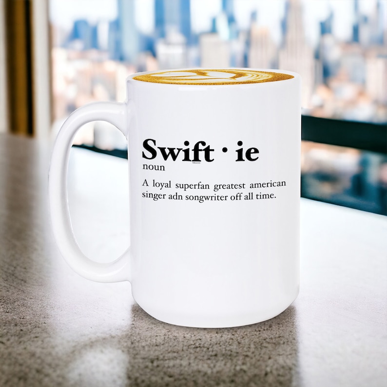 Pu&ograve; includere: Tazza in ceramica bianca con la parola "Swiftie" in nero e una definizione. La tazza contiene un latte con un design decorativo. Lo sfondo mostra un paesaggio urbano sfocato.