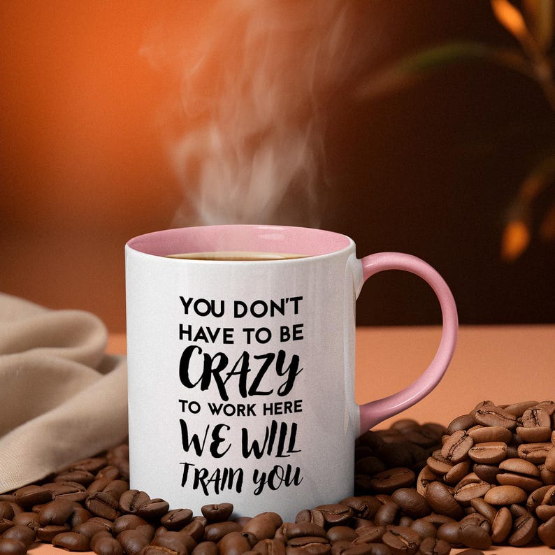 K&ouml;nnte beinhalten: Eine wei&szlig;e Kaffeetasse mit rosafarbenem Rand und Henkel. Die Tasse hat einen schwarzen Text mit der Aufschrift "You don't have to be crazy to work here we will train you".