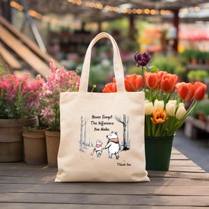 May include: Cream-colored tote bag featuring a graphic of a bear and pig walking together in a forest scene. The text on the bag reads "Never Forget The Difference You Make. Thank You."