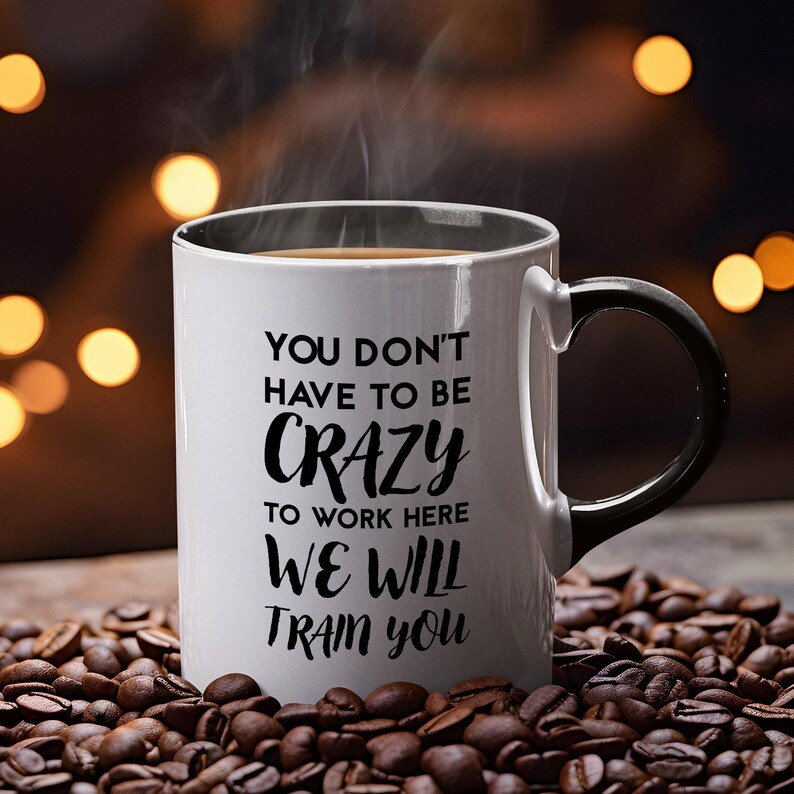 K&ouml;nnte beinhalten: Eine wei&szlig;e Kaffeetasse mit schwarzem Henkel. Die Tasse hat einen schwarzen Text mit der Aufschrift "You don't have to be crazy to work here, we will train you."