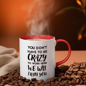K&ouml;nnte beinhalten: Eine wei&szlig;e Keramik-Kaffeetasse mit rotem Rand und Henkel. Die Tasse hat einen schwarzen Text mit der Aufschrift "You don't have to be crazy to work here we will train you". Die Tasse steht auf einem Haufen Kaffeebohnen.
