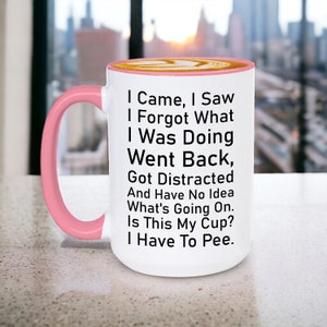 May include: White ceramic coffee mug with a pink rim and handle. The mug has black text that reads: "I Came, I Saw, I Forgot What I Was Doing, Went Back, Got Distracted And Have No Idea What's Going On. Is This My Cup? I Have To Pee."