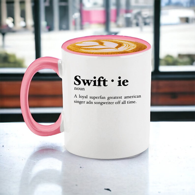 Pu&ograve; includere: Tazza da caff&egrave; bianca con manico e bordo rosa, riempita con un latte. La tazza presenta il testo "Swiftie" con una definizione: "Una superfan leale, la pi&ugrave; grande cantante e cantautrice americana di tutti i tempi."
