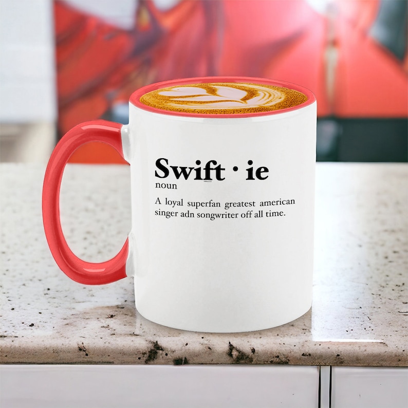 Pu&ograve; includere: Tazza da caff&egrave; bianca con manico e bordo rossi, piena di caff&egrave;. La tazza presenta la parola "Swiftie" e una definizione. La definizione recita: "Una superfan leale, la pi&ugrave; grande cantante e cantautrice americana di tutti i tempi."